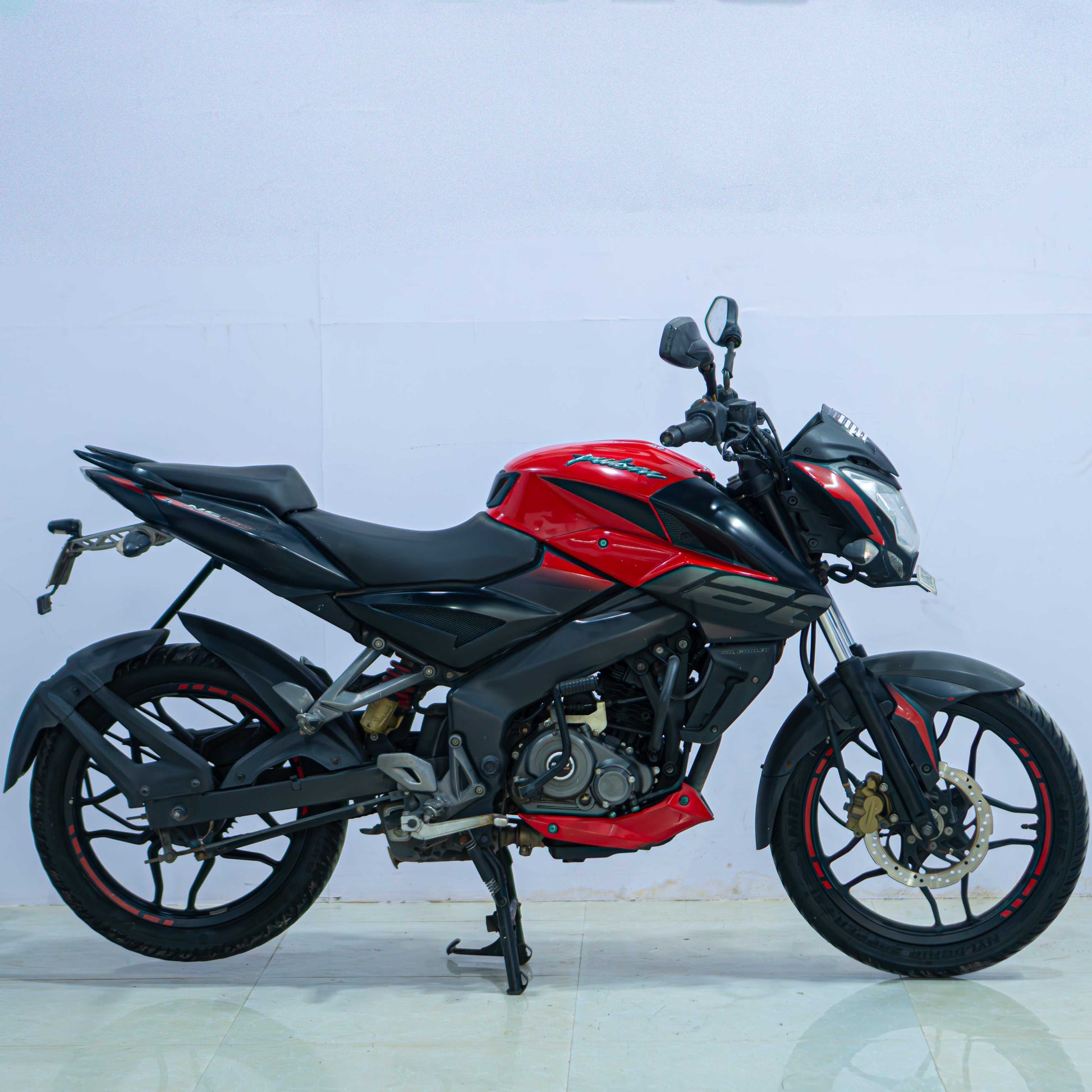 Red Bajaj NS 160 motorcycle parked on a city street, showcasing its sporty design and sleek bodywork.