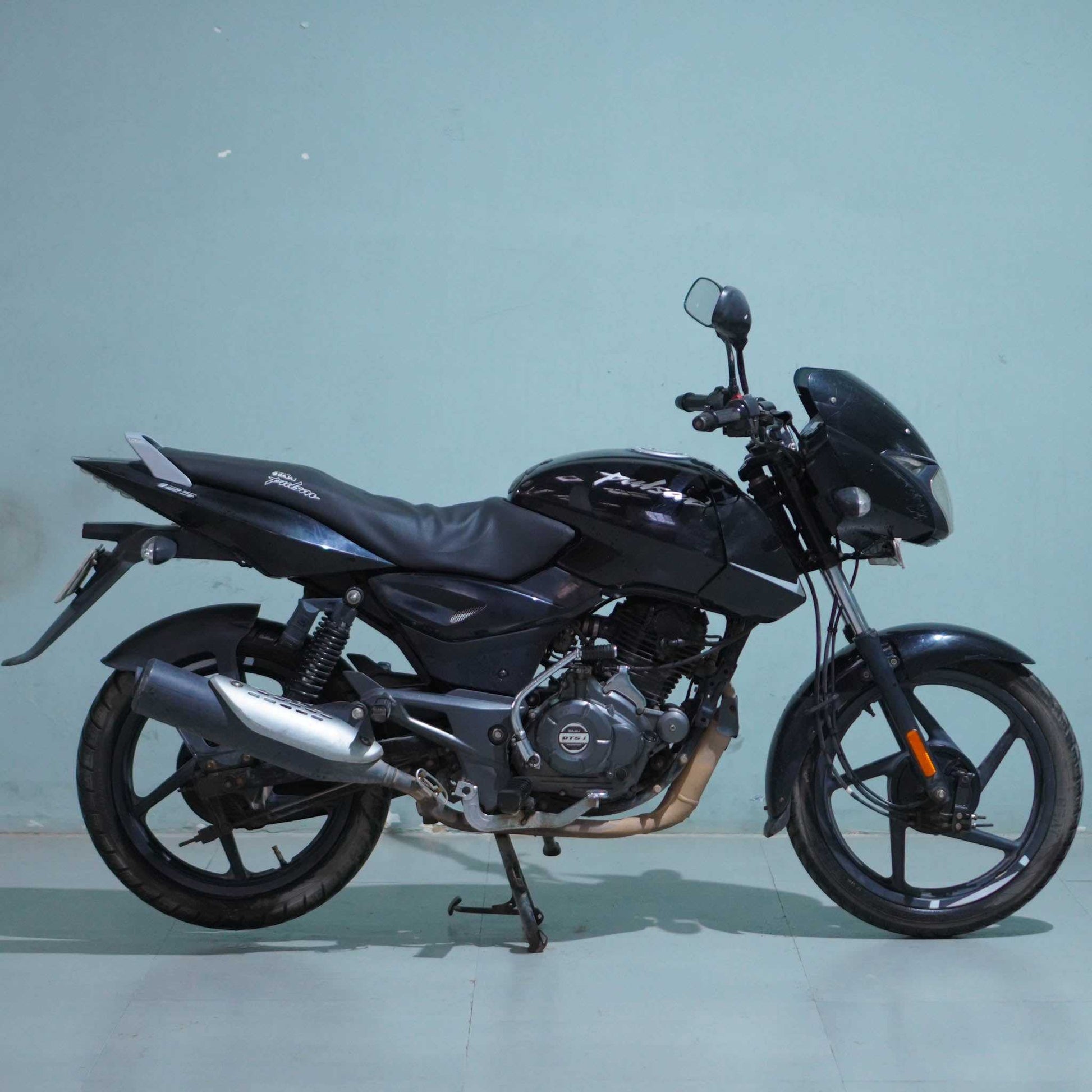 Red Bajaj Pulsar 125 motorcycle parked on a street, featuring a sporty design and sleek body with brand logo visible.